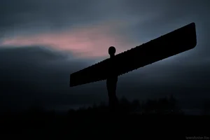 The Angel of the North sculpture silhouetted against a dark, cloudy sky with a faint glow of evening light behind it.