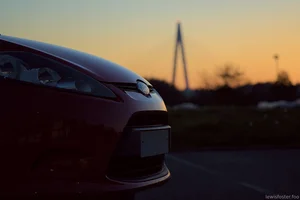 A close-up of the front corner of a red car at sunset, with a softly blurred bridge silhouetted against a warm evening sky in the background.
