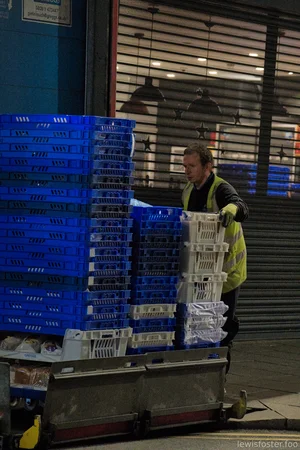 A worker in a high-visibility vest unloads stacked blue and white plastic crates from a Greggs delivery truck in the evening.