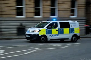 A police van with blue lights flashing drives through a city street, motion-blurred against a stone building as it passes an urban intersection.